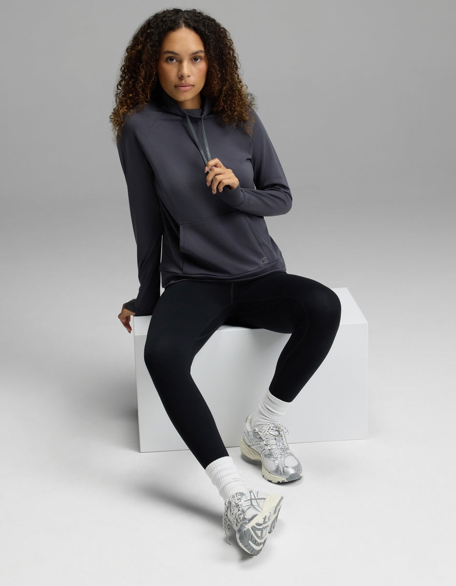 A person with curly hair wearing the Cozy Earth Women’s Brushed Bamboo Mock Neck, paired with black leggings and silver sneakers, sits on a white block in a studio with a plain gray background.|Color:Graphite