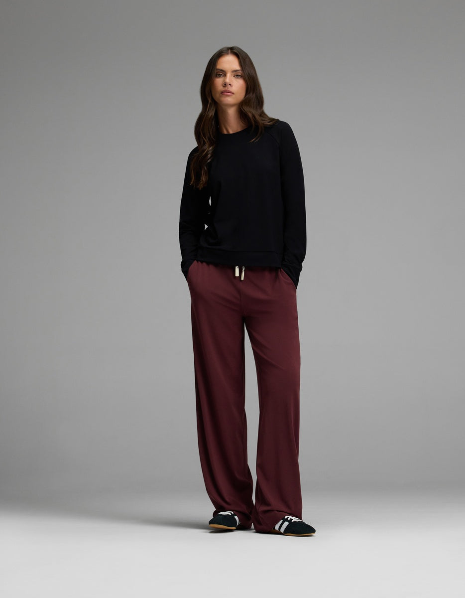 A woman stands with her hands in her pockets, wearing the Cozy Earth Women’s Brushed Bamboo Pullover Crew in black, loose burgundy pants, and black Adidas slides against a plain gray background.|Color:Black