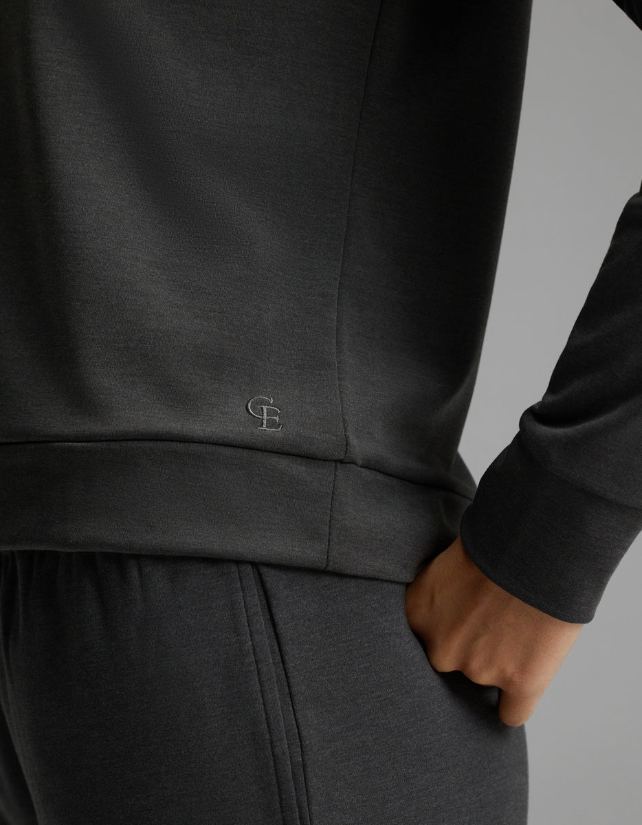 A person is wearing the Cozy Earth Women’s Brushed Bamboo Pullover Crew and matching pants, with one hand in the pocket. The dark gray sweatshirt features small “CE” initials stitched near the hem, set against a plain gray background.|Color:Charcoal