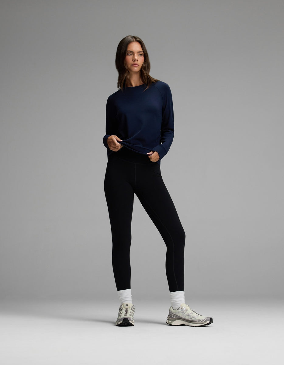 A woman stands against a plain gray background, wearing the Cozy Earth Women’s Brushed Bamboo Pullover Crew in navy, paired with black leggings, white socks, and white sneakers. She holds the hem of her pullover and looks slightly to the side with a neutral expression.|Color:Navy