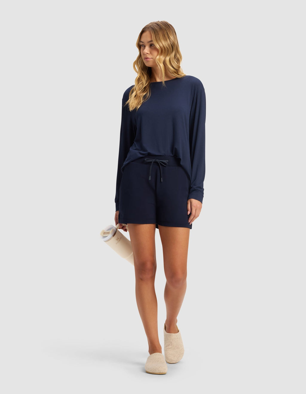 A woman with long wavy hair wears Cozy Earth’s Women’s Brushed Bamboo Shorts in navy blue with a matching long-sleeve top, holds a rolled-up towel, and dons beige slippers against a plain light gray background.|Color:Navy