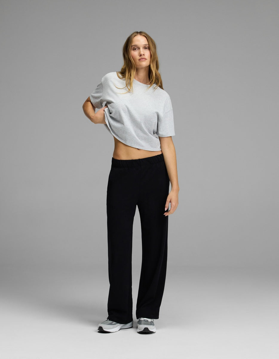 A person stands against a plain gray background, wearing a light gray t-shirt, gray sneakers, and Cozy Earth’s Women’s Brushed Bamboo Wide Leg Pull-On Pant in black. One hand rests on their hip while the other arm hangs loosely by their side.|Color:Black