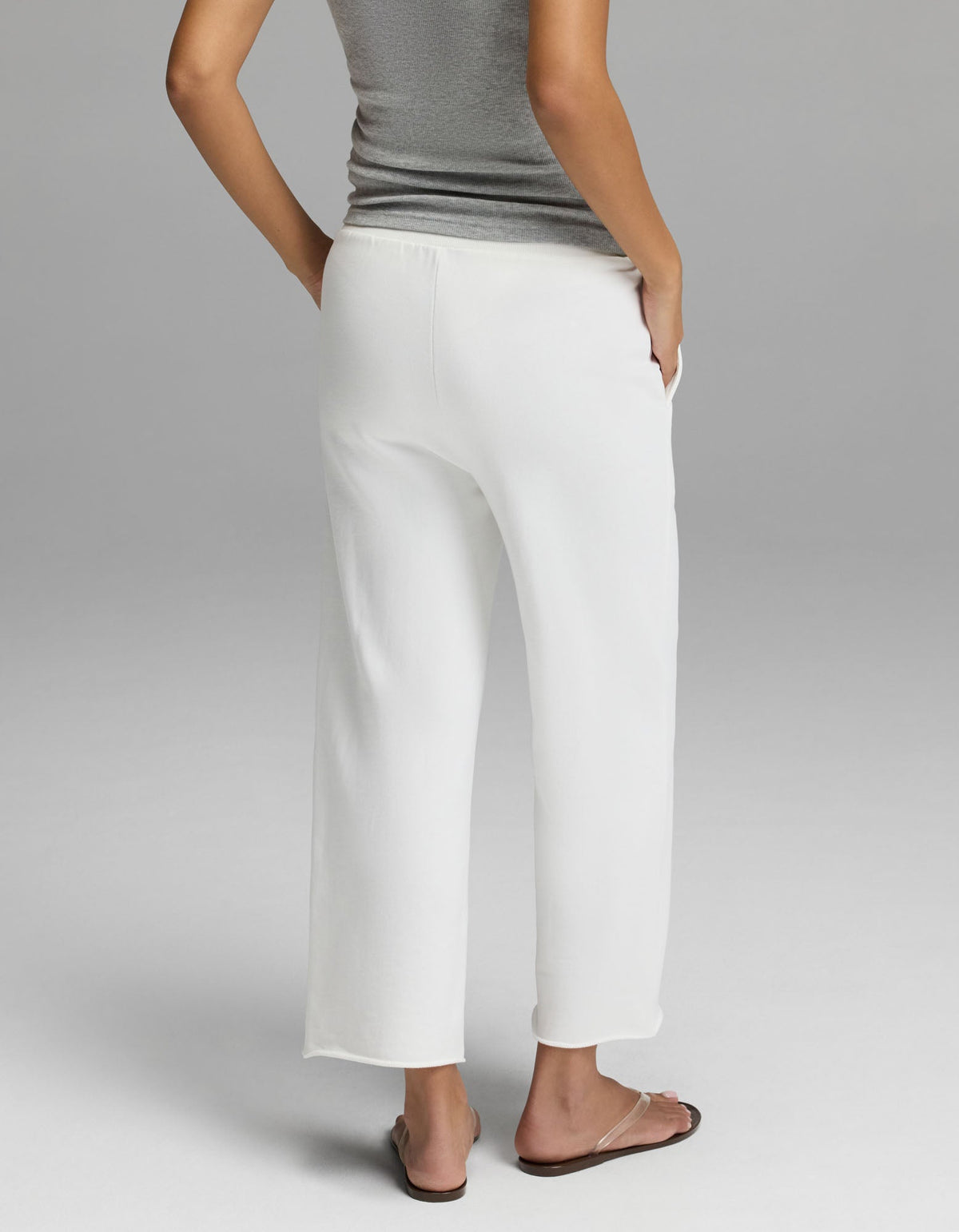 A woman is photographed from behind against a plain light gray background, standing with one hand in the pocket of her loose white Cozy Earth Women’s Chelsea Cropped Pant, paired with a gray sleeveless top and flip-flops.|Color:Bone