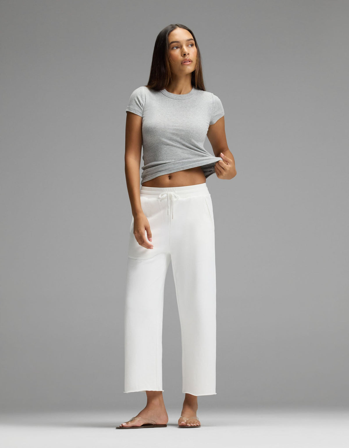 A woman wears the Cozy Earth Women’s Chelsea Cropped Pant, standing barefoot on a gray background in a gray t-shirt, lifting one side of her shirt and gazing slightly upward.|Color:Bone