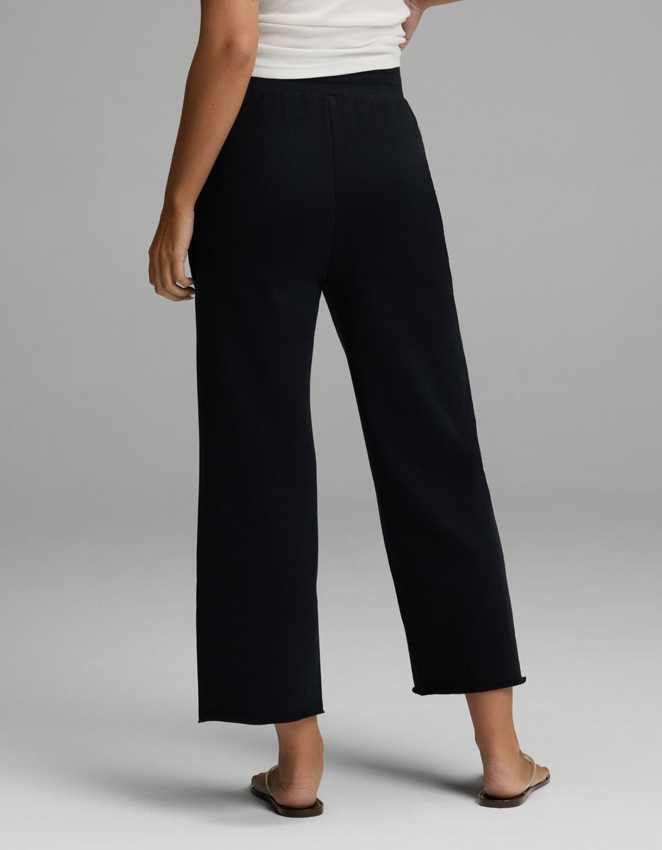 A person is shown from the back standing on a light gray background, wearing Cozy Earth’s Women’s Chelsea Cropped Pant in black, paired with a white sleeveless top and beige flat sandals.|Color:Jet Black