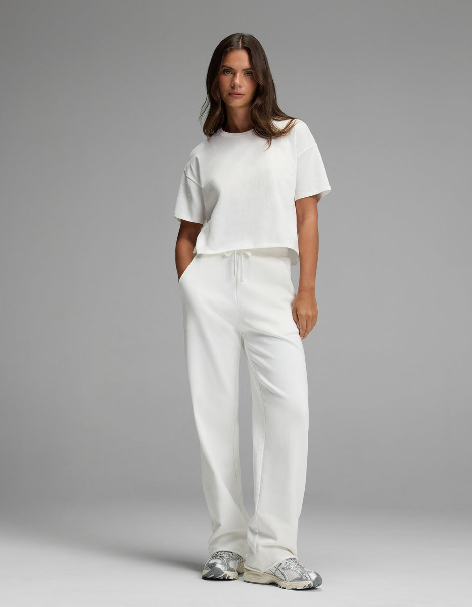A woman stands against a gray background with her hands in her pockets and a neutral expression, wearing Cozy Earth’s Women’s Chelsea Pant in white paired with a white t-shirt and silver sneakers.|Color:Bone