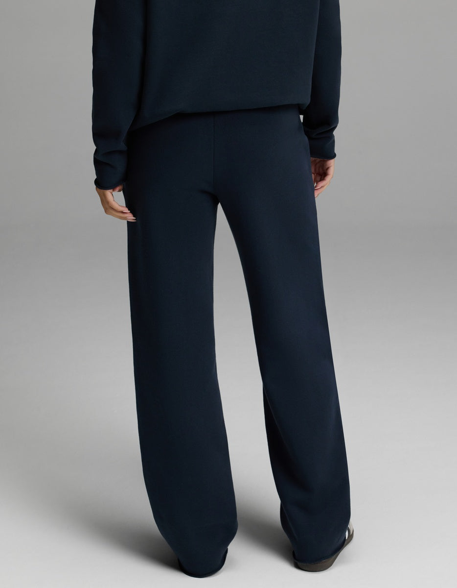 A person is pictured from behind, indoors against a plain light gray background, wearing Cozy Earth’s Women’s Chelsea Pant paired with a dark navy sweater. The lower half of the body is visible.|Color:Eclipse