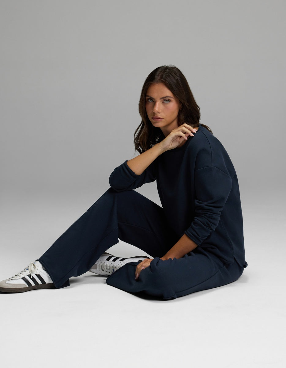 A person with long brown hair sits on the floor in a relaxed pose, wearing the Cozy Earth Women’s Chelsea Pant in navy blue, paired with a matching sweatshirt and white sneakers featuring black stripes. The background is plain and gray.|Color:Eclipse
