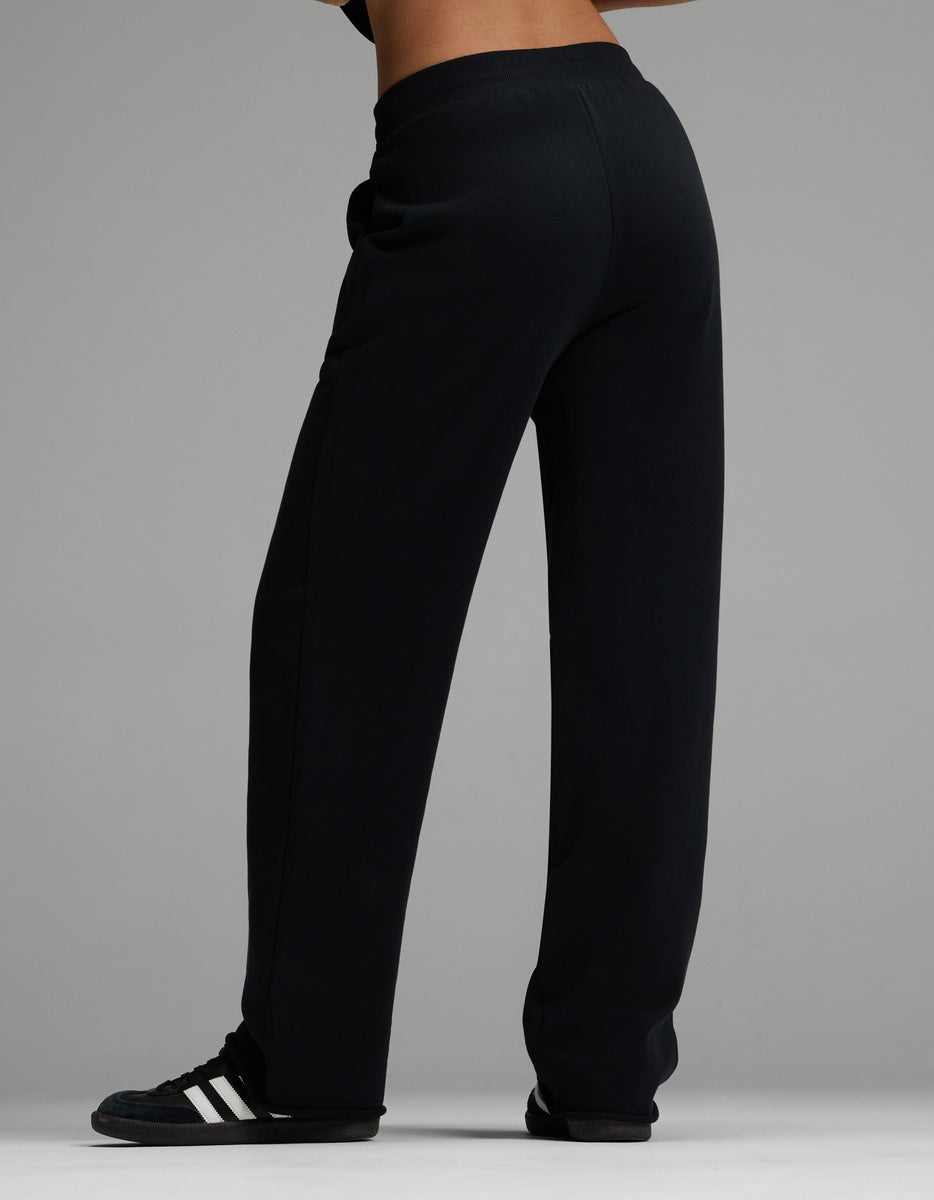 A woman is shown from behind against a gray background, wearing Cozy Earth’s Women’s Chelsea Pant in black and black Adidas sneakers with white stripes, with the image focused on the lower half of her body.|Color:Jet Black