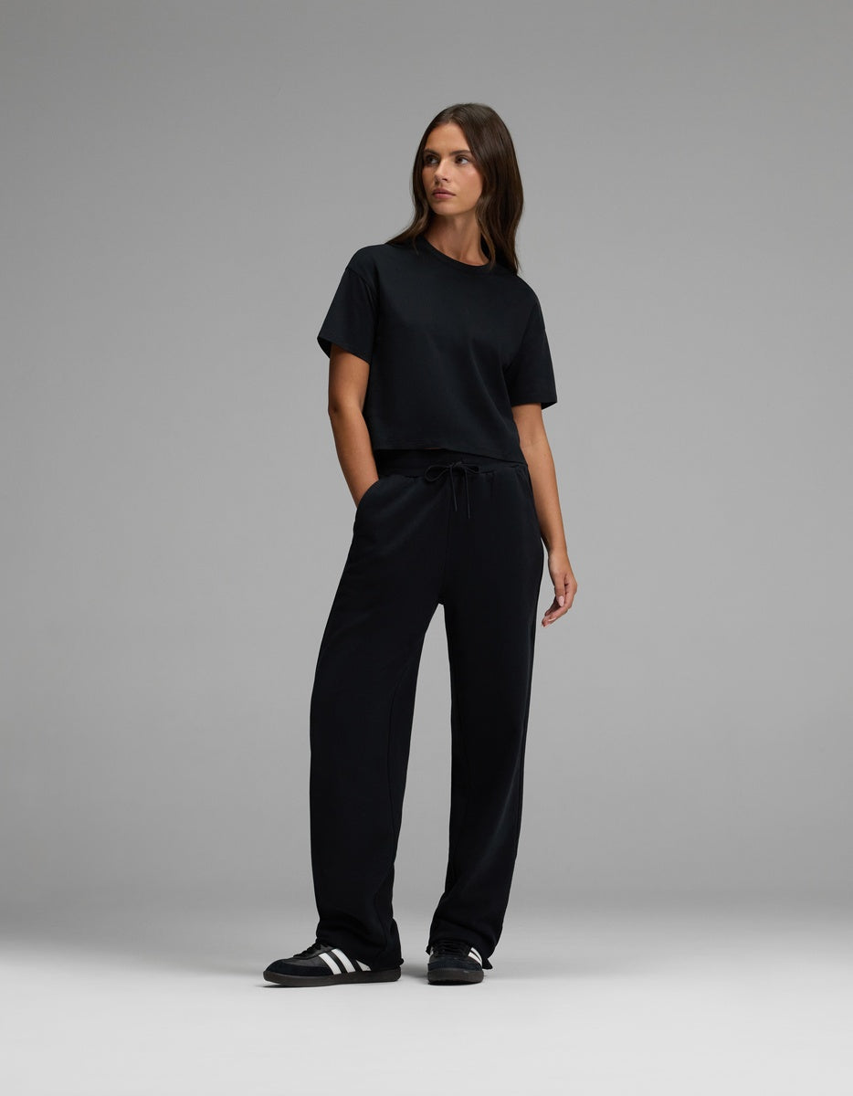 A woman stands against a gray background, wearing black Cozy Earth Women’s Chelsea Pants, a black t-shirt, and black Adidas sneakers with white stripes. She has her hands in her pockets and looks slightly to her left.|Color:Jet Black
