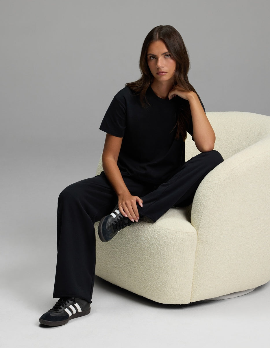 A woman with long brown hair, wearing a black t-shirt, black sneakers, and the Cozy Earth Women’s Chelsea Pant, sits on a cream textured armchair against a plain gray background.|Color:Jet Black