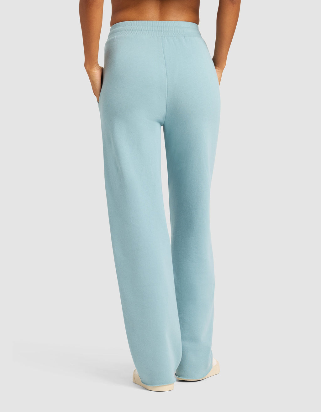 Model wearing Cozy Earth Chelsea Pant in light blue, paired with white shoes. Shown from the back with hands in pockets against a plain light background.|Color:River