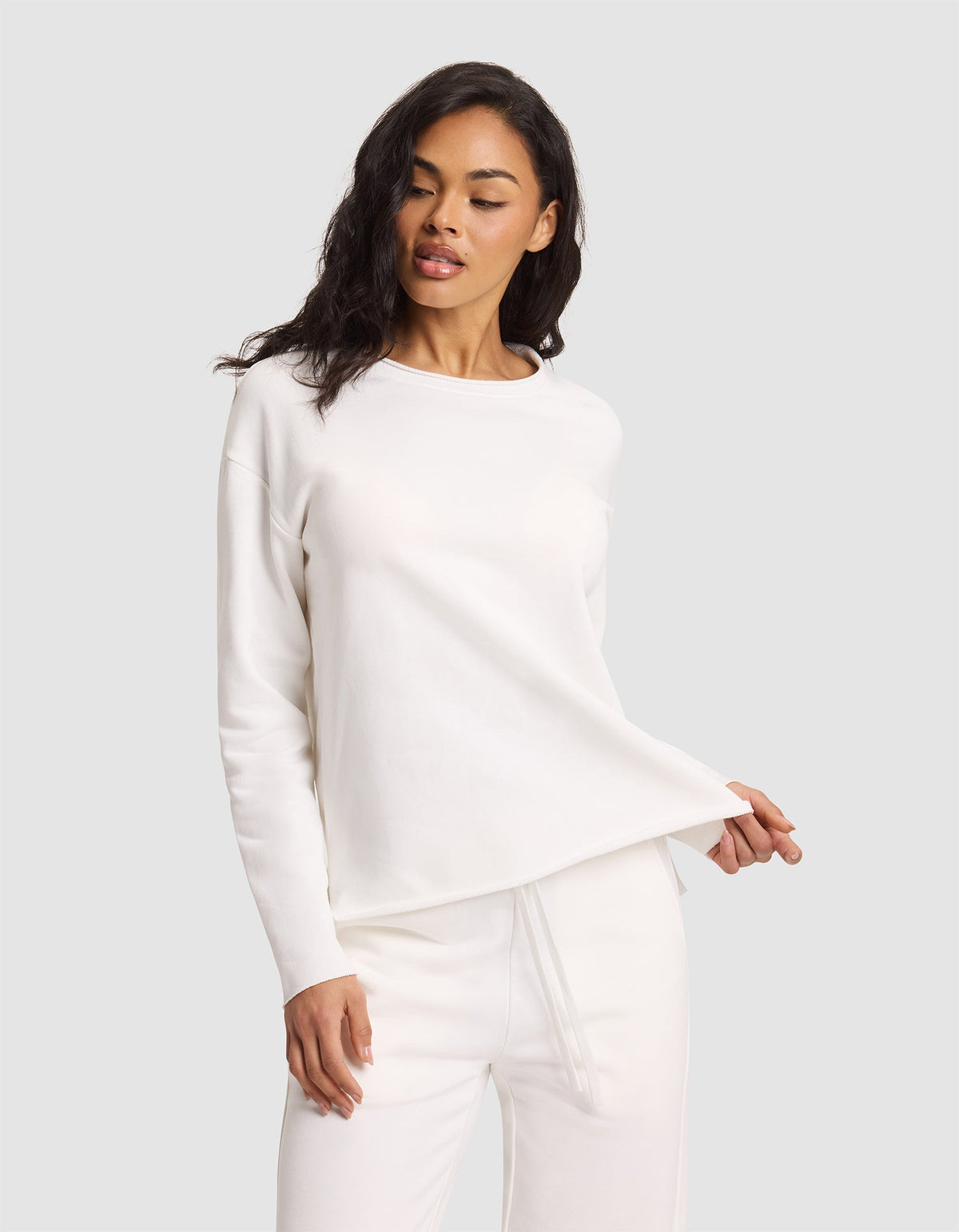 A woman poses against a light gray background wearing the Cozy Earth Women's Chelsea Pullover and matching pants in all white, holding the hem of the top with one hand while looking slightly downward. |Color:Bone