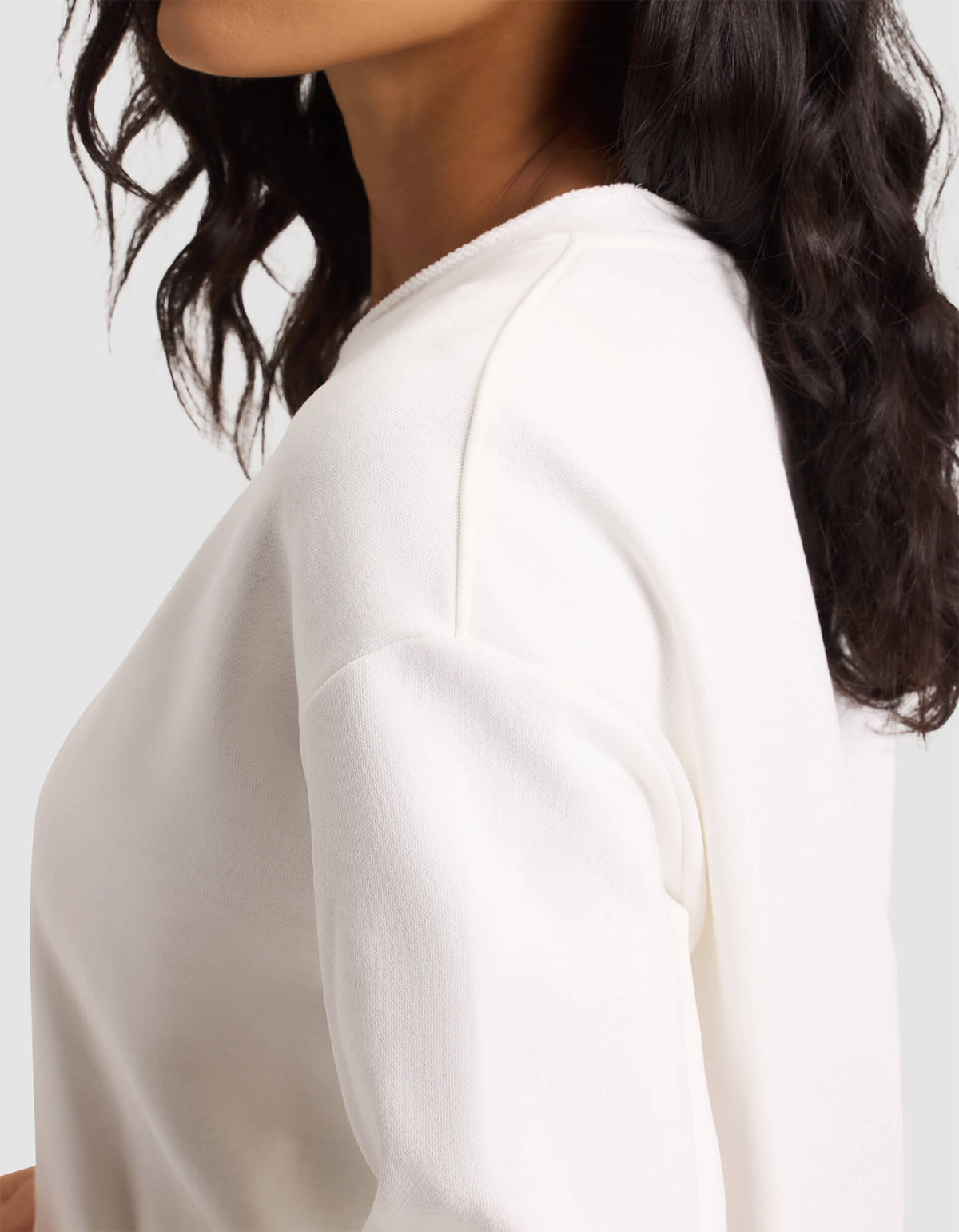 A woman with wavy dark hair wears the Cozy Earth Women's Chelsea Pullover, an off-white long-sleeve top, shown from the shoulders up and slightly turned to the side against a plain light background. |Color:Bone