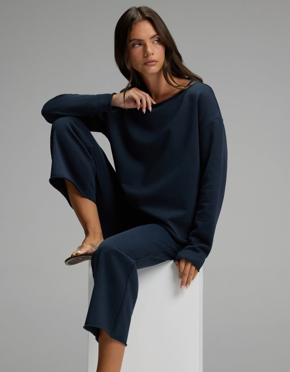Wearing the Cozy Earth Women’s Chelsea Cropped Set, a woman sits on a white block with one knee raised and the other leg hanging down, gazing to the side against a plain gray background.|Color:Eclipse