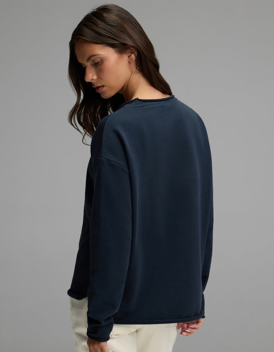 A woman with long brown hair stands against a plain gray background, looking over her shoulder while wearing the Cozy Earth Women’s Chelsea Pullover in dark blue and light pants.|Color:Eclipse