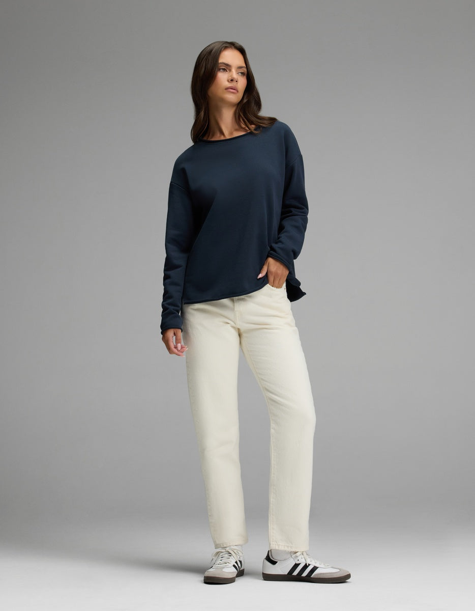 A woman stands against a gray background, wearing Cozy Earth’s Women’s Chelsea Pullover in navy blue, paired with off-white pants and white adidas sneakers with black stripes. She has straight brown hair and a neutral expression, with one hand in her pocket.|Color:Eclipse