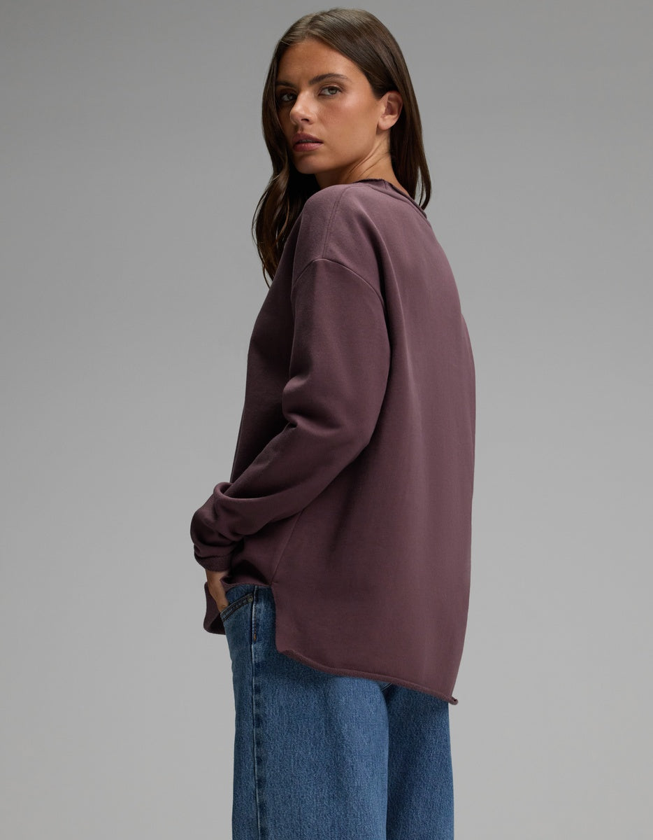 A person with long brown hair stands sideways and looks over their shoulder against a plain gray background, wearing the Cozy Earth Women’s Chelsea Pullover in purple paired with blue jeans.|Color:Fig
