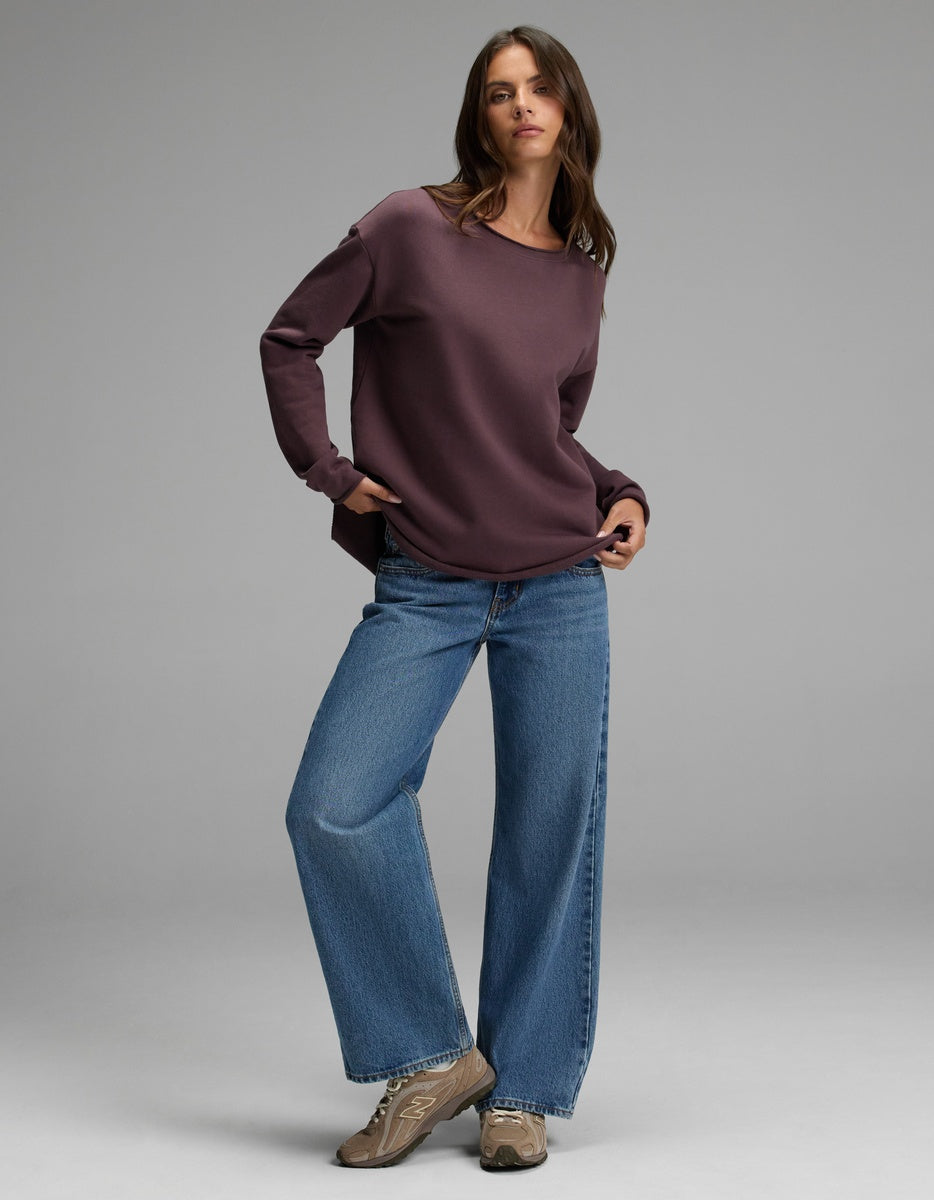 A woman with long brown hair and a relaxed expression poses in a simple studio, wearing the Cozy Earth Women’s Chelsea Pullover—a loose, dark purple sweatshirt—paired with wide-leg blue jeans and tan sneakers.|Color:Fig
