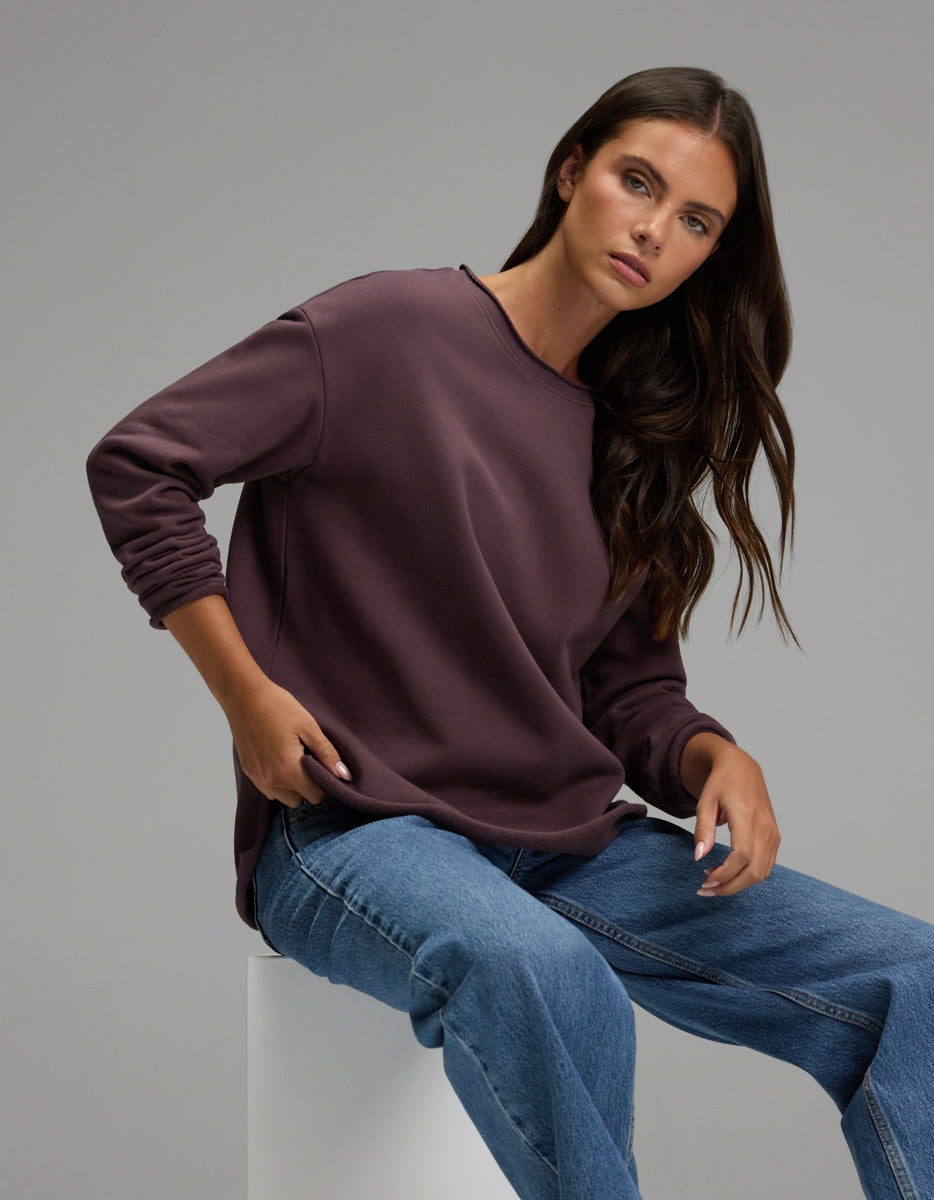 A woman with long brown hair sits on a white block against a plain gray background, looking at the camera with a relaxed expression while wearing the Cozy Earth Women’s Chelsea Pullover in maroon and blue jeans.|Color:Fig