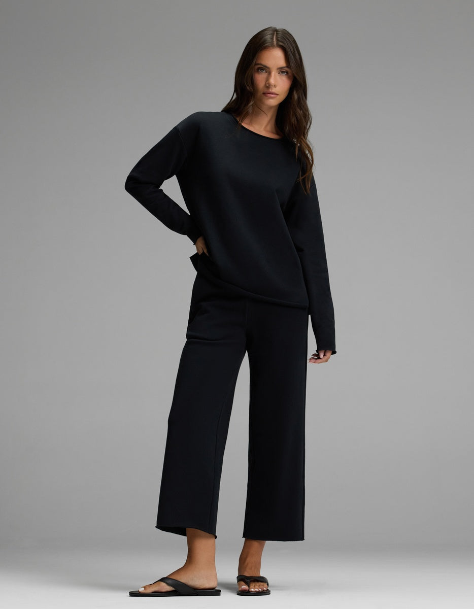 A woman with long brown hair and a relaxed expression poses against a gray backdrop, wearing the Cozy Earth Women’s Chelsea Pullover in black, paired with wide-leg cropped pants and black sandals. She has one hand in her pocket.|Color:Jet Black