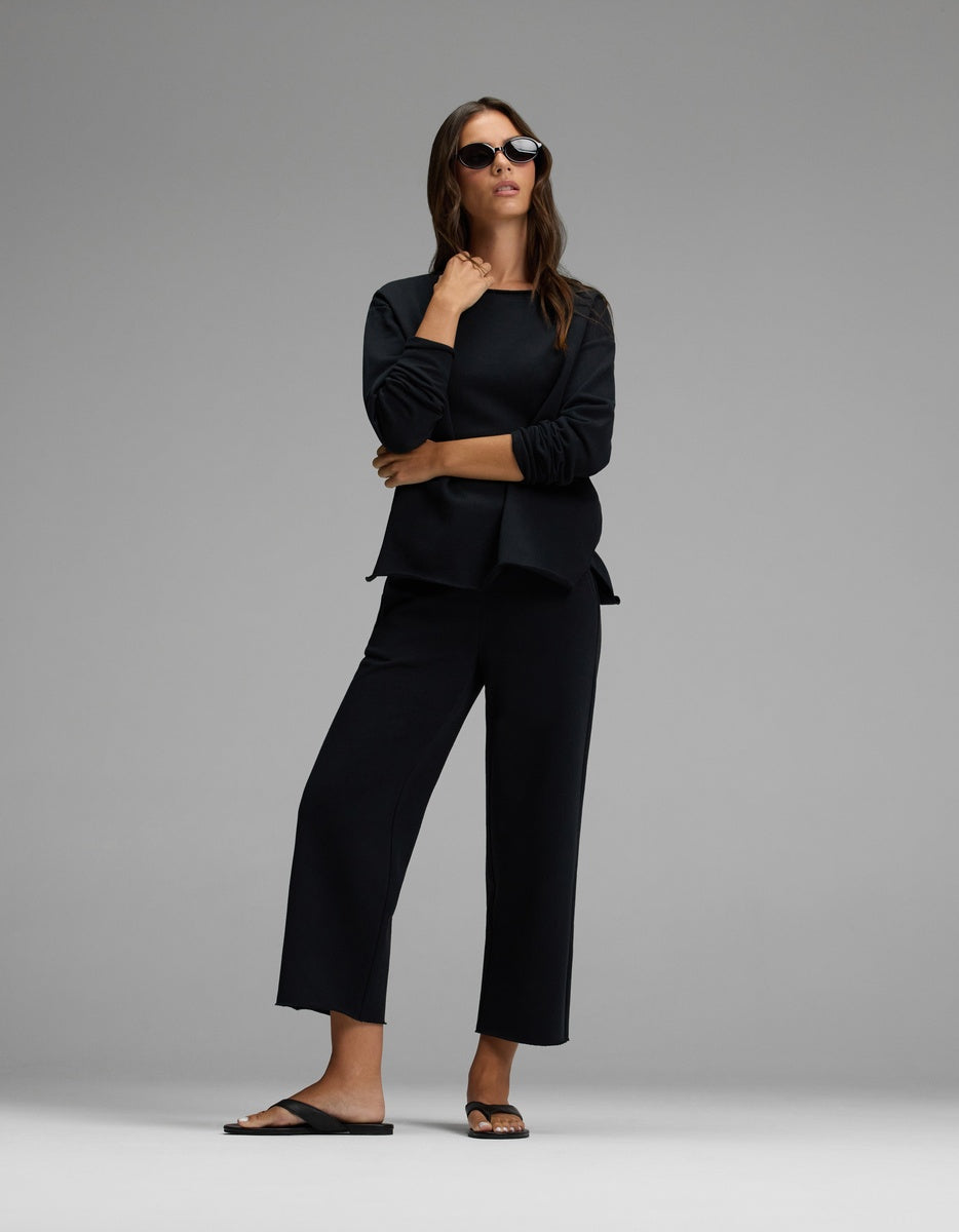A woman stands confidently against a plain gray background, wearing the Cozy Earth Women’s Chelsea Pullover in black, paired with black wide-leg pants, black sunglasses, and black sandals.|Color:Jet Black