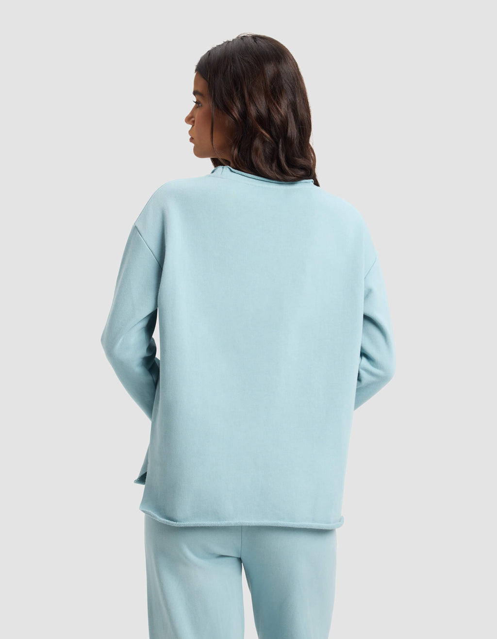 A woman with long, wavy dark hair is seen from behind wearing the Cozy Earth Women's Chelsea Pullover in light blue against a plain backdrop.|Color:River