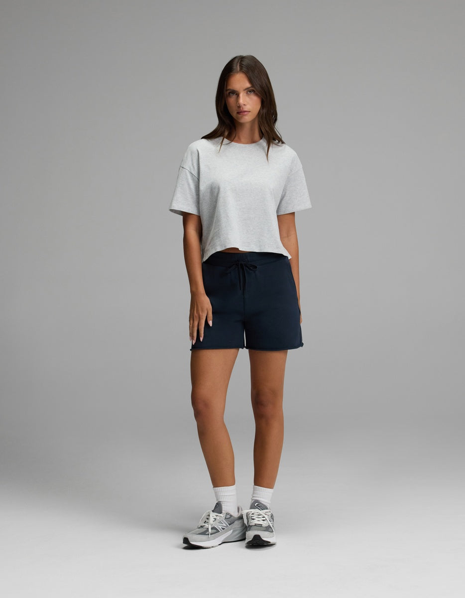 A woman stands against a plain background, wearing Cozy Earth’s Women’s Chelsea Short, paired with a light gray T-shirt and gray sneakers. She has long brown hair and a neutral expression, with her hands relaxed by her sides.|Color:Eclipse