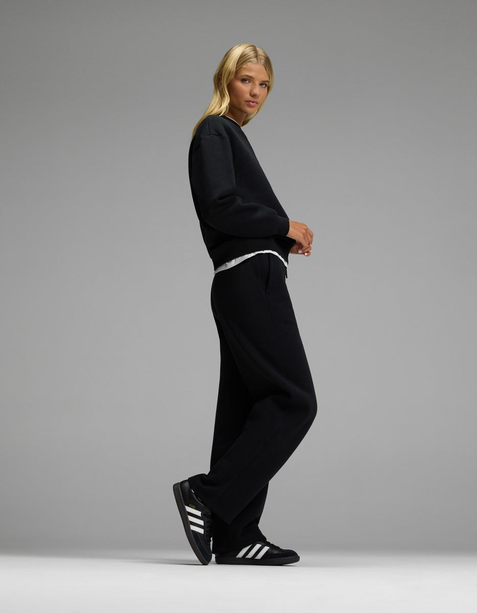 A person with long blonde hair wears Cozy Earth’s Women’s CityScape Crewneck & Wide Leg Pant Set in black, paired with black Adidas sneakers featuring white stripes, standing against a plain gray backdrop.|Color:Jet Black
