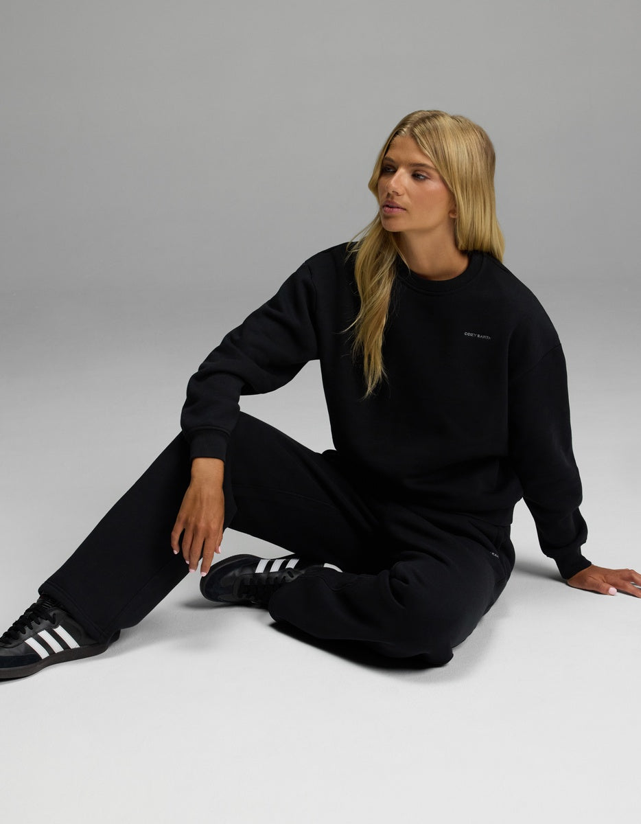 A person with long blonde hair sits on the floor wearing the Cozy Earth Women’s CityScape Crewneck & Wide Leg Pant Set in black, looking to the side against a plain light gray background.|Color:Jet Black