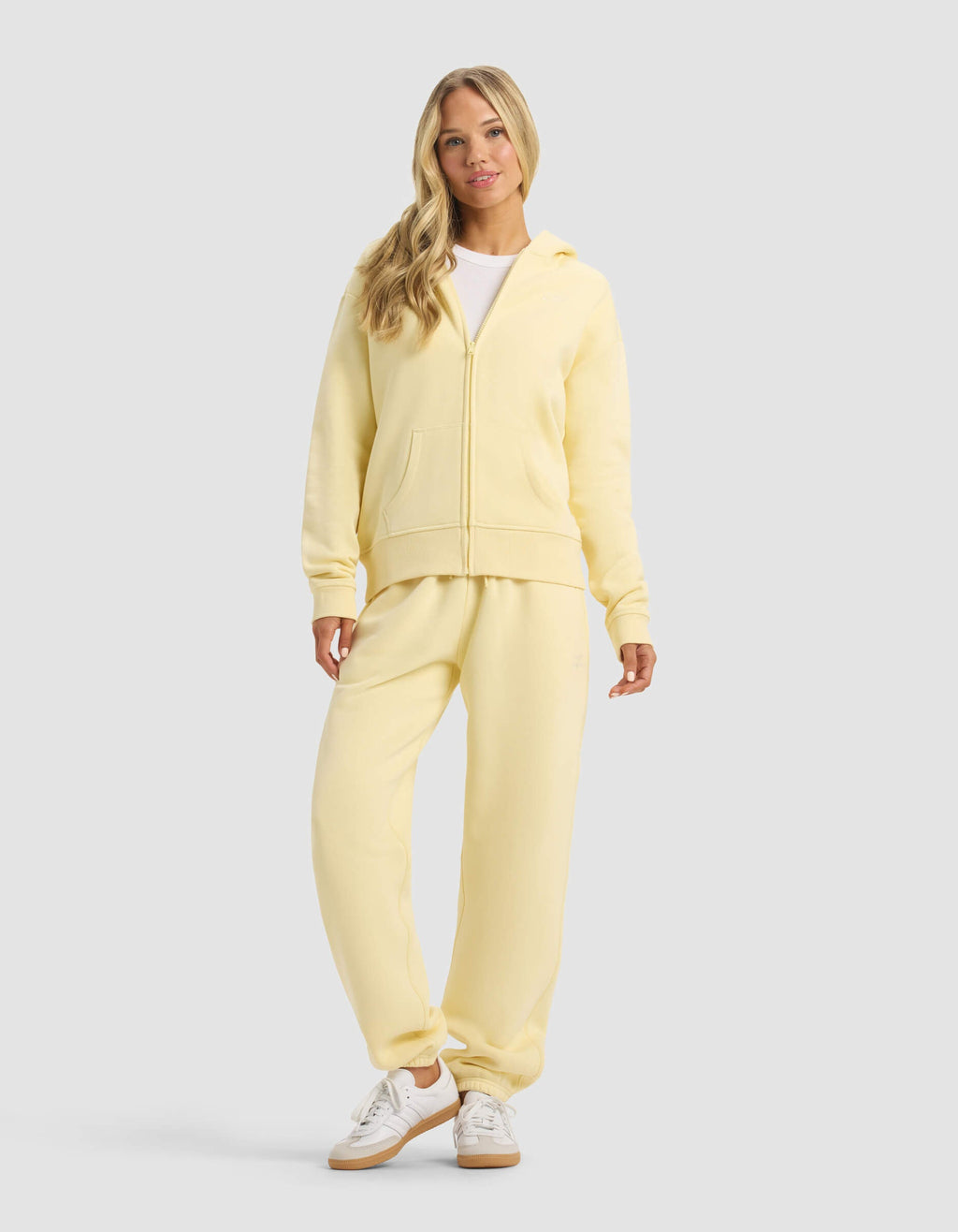 A woman with long blonde hair smiles softly against a gray background, wearing the Cozy Earth Women's CityScape Full Zip hoodie in light yellow, matching sweatpants, a white shirt, and white sneakers.|Color:Meringue