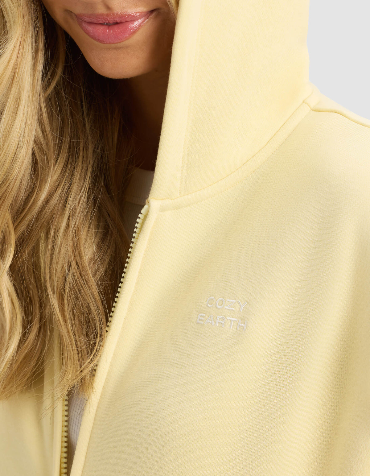 A close-up shows a woman wearing the Cozy Earth Women's CityScape Full Zip hoodie in light yellow, with the hood up and only her lower face and blond hair visible. "COZY EARTH" is embroidered in white on the chest.|Color:Meringue
