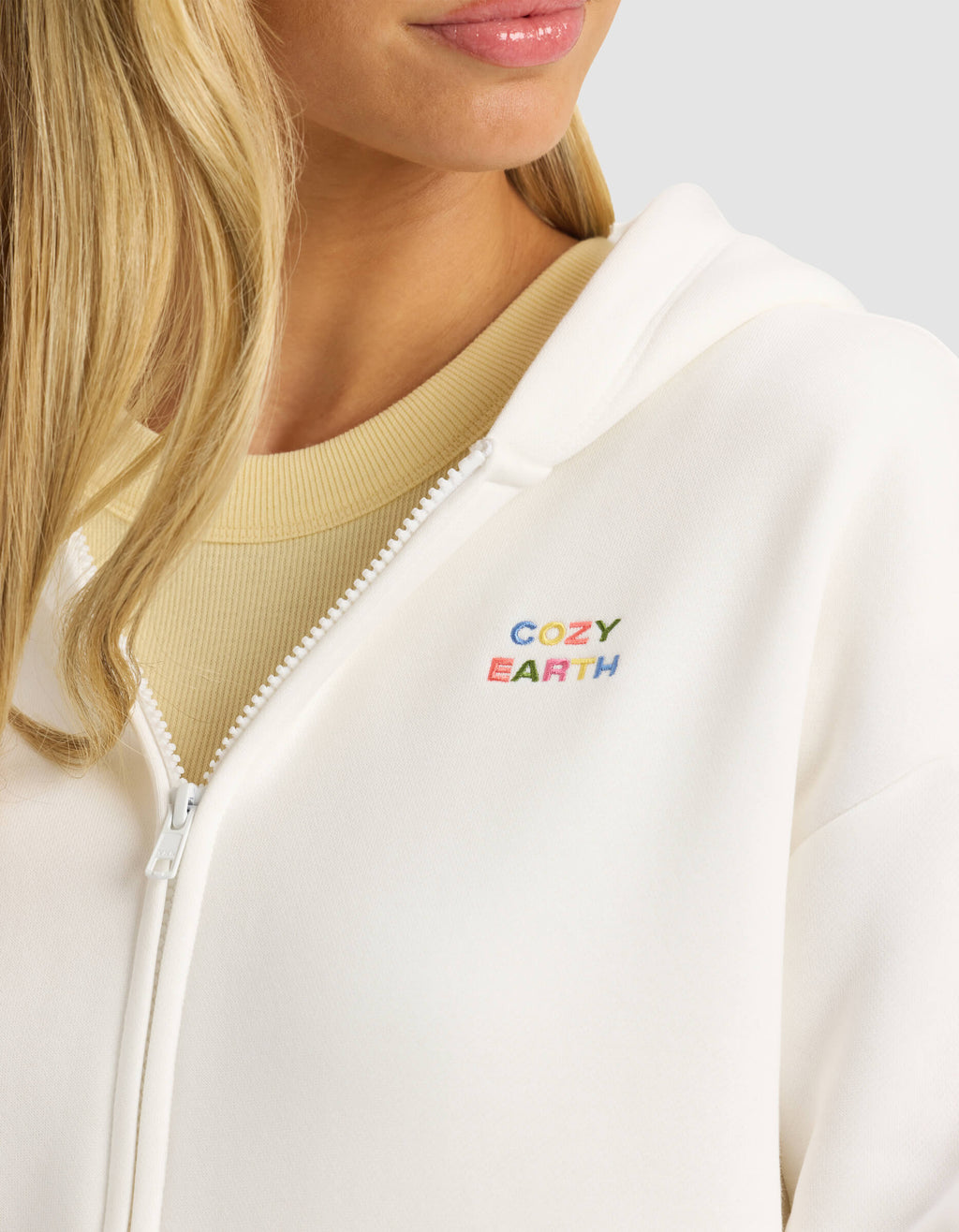 A woman wears the Cozy Earth Women's CityScape Full Zip—a white hoodie with colorful "COZY EARTH" embroidery on the chest—over a beige top. Only her lower face, neck, and part of her blonde hair are visible.|Color:Sea Salt