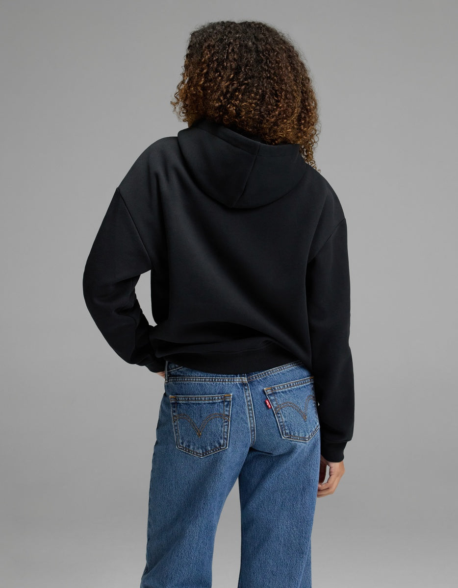 A woman with curly hair is seen from behind, wearing the Cozy Earth Women’s CityScape Hoodie in black and blue jeans, standing against a plain gray backdrop.|Color:Jet Black