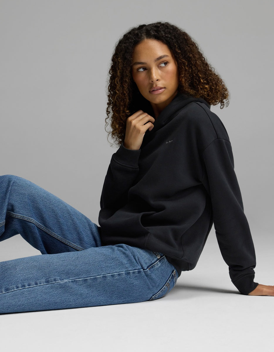 A woman with curly hair sits on the floor, wearing the Cozy Earth Women’s CityScape Hoodie and blue jeans, looking to the side against a plain gray background.|Color:Jet Black