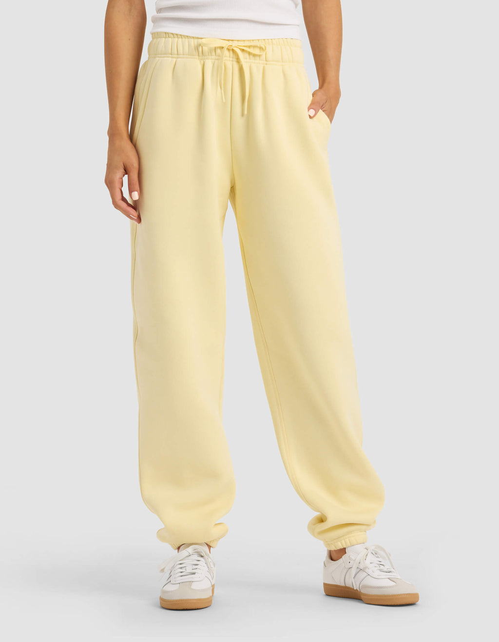 A person stands against a plain background with hands in the pockets of Cozy Earth Women's Cityscape Sweatpant in light yellow, paired with a white top and white sneakers.|Color:Meringue