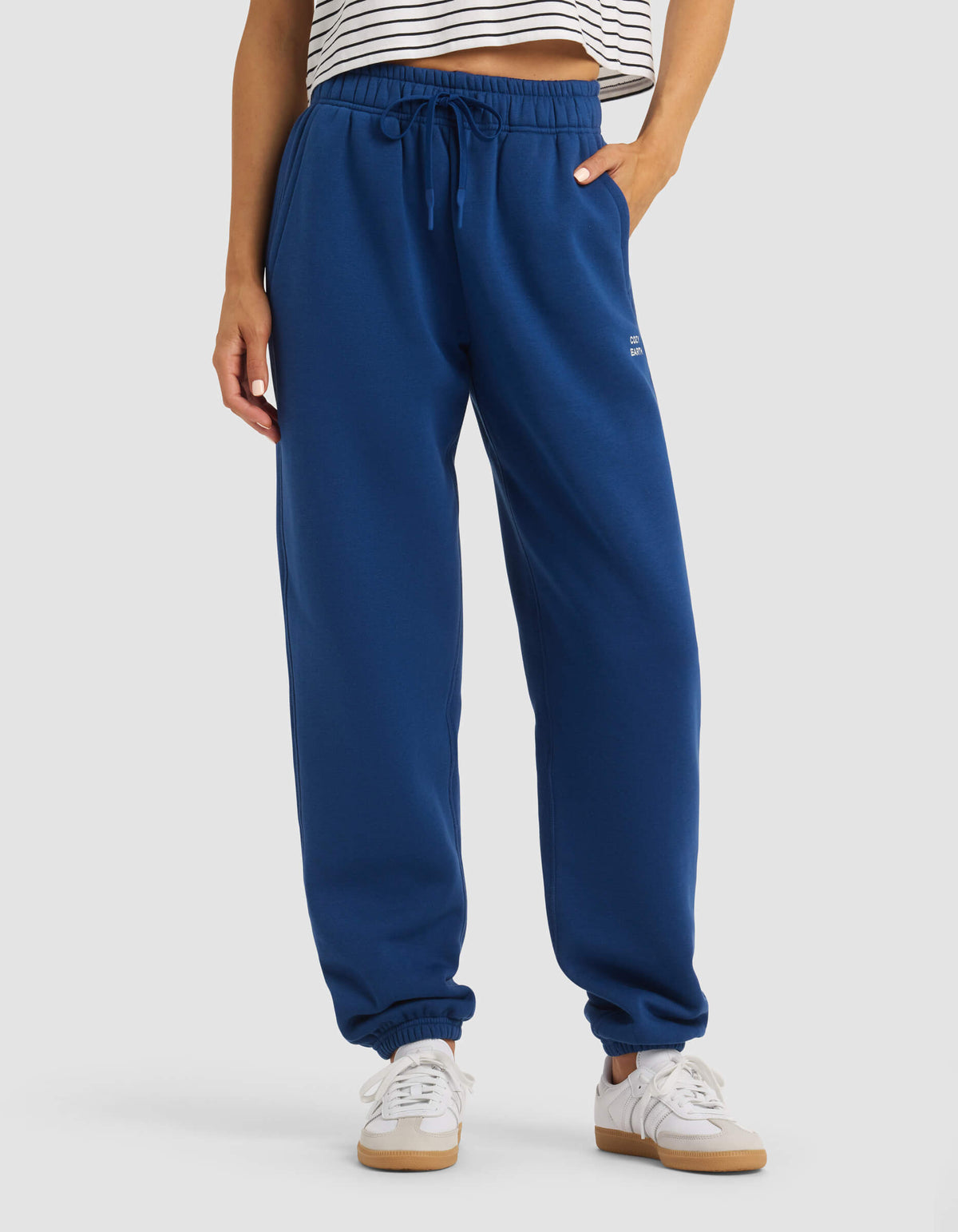 A person wearing Cozy Earth's Women's Cityscape Sweatpant in royal blue, paired with a striped crop top and white sneakers, stands against a plain background, showing only their lower torso and legs.|Color:Pacific