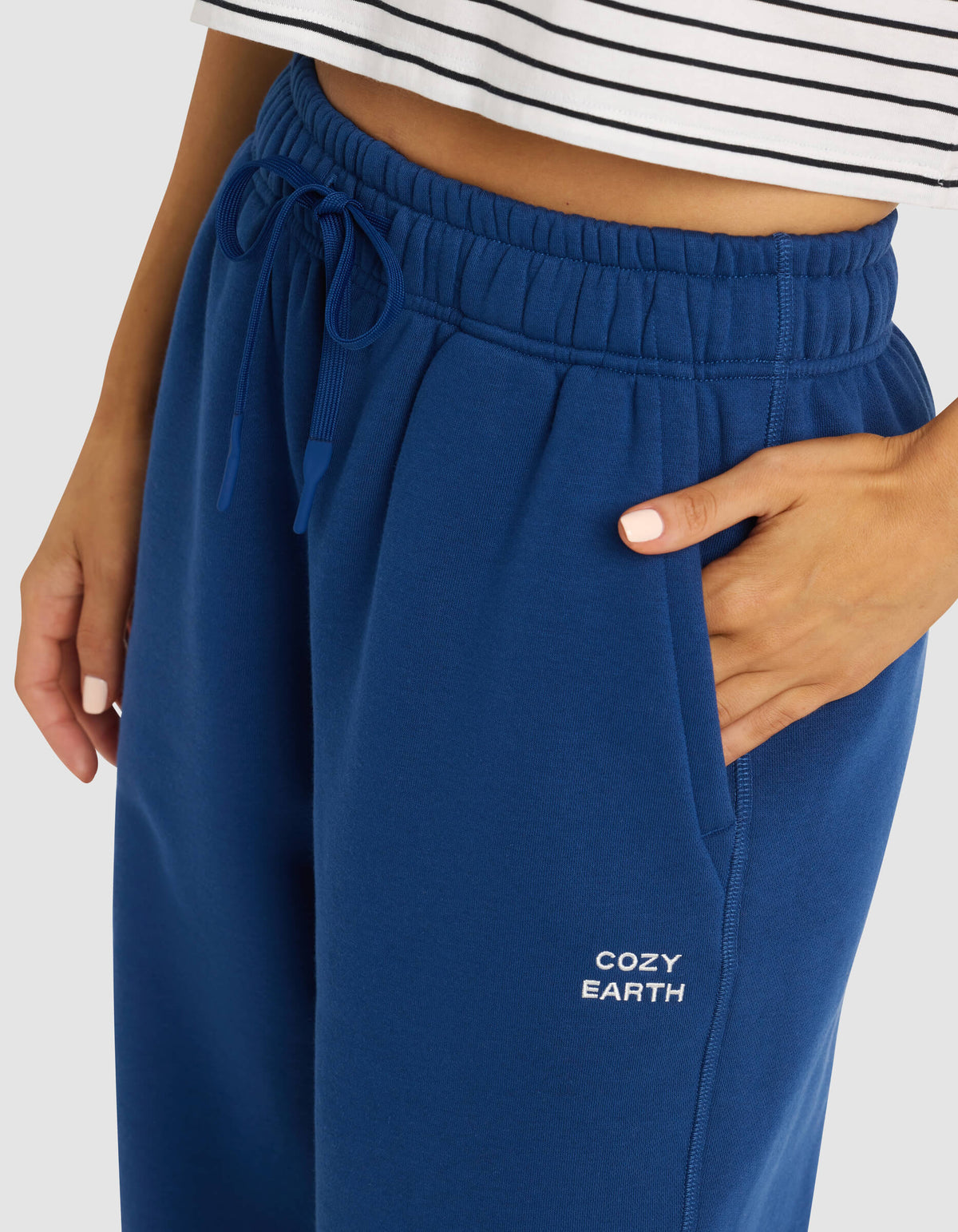 A woman wears Cozy Earth's Women's Cityscape Sweatpant in blue, featuring the brand name on the pocket, paired with a white striped crop top and one hand in her pocket.|Color:Pacific