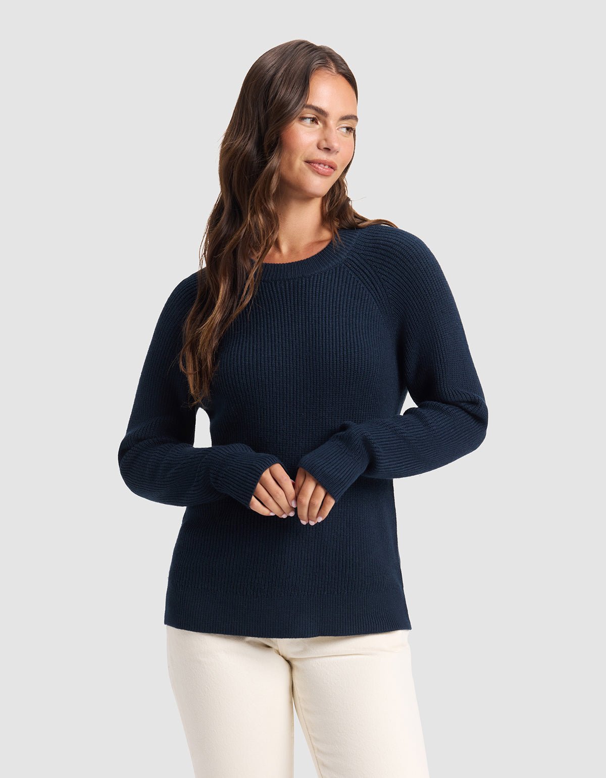 A woman with long brown hair wears the Cozy Earth Women's Classic Crewneck in navy blue and cream-colored pants, standing against a light gray background and smiling gently while looking to the side.|Color:Eclipse