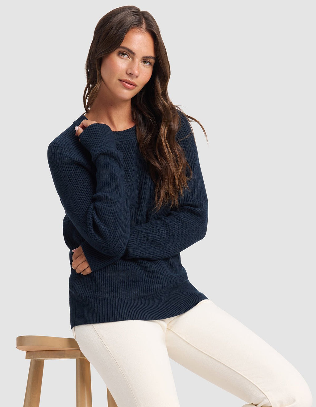 A woman with long brown hair wears the Cozy Earth Women's Classic Crewneck in navy blue and off-white pants, seated on a wooden stool against a plain light gray background.|Color:Eclipse