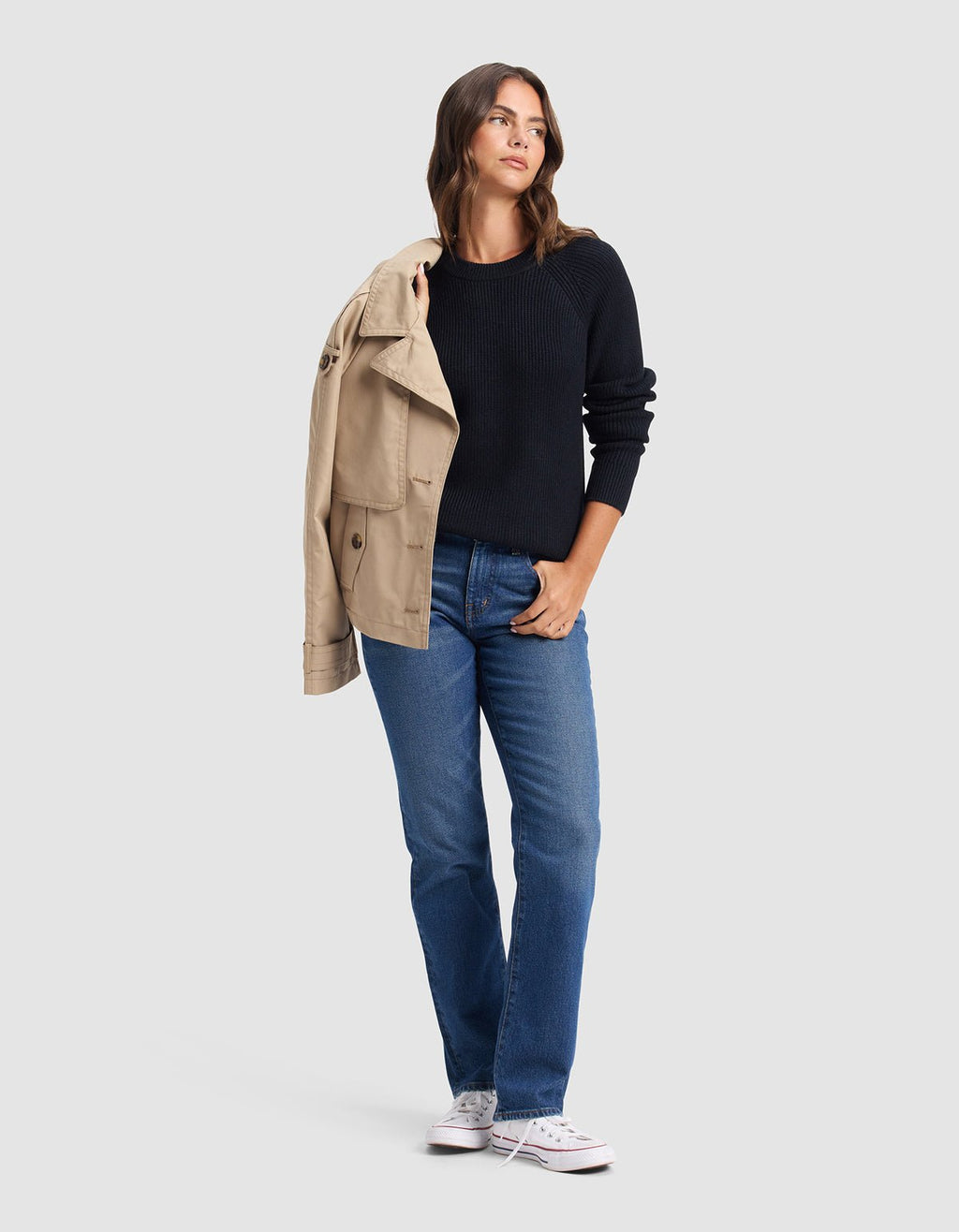 A woman stands against a plain background, wearing Cozy Earth's Women's Classic Crewneck in black, styled with blue jeans and white sneakers. She drapes a tan coat over one shoulder and glances to the side with one hand in her pocket.|Color:Jet Black
