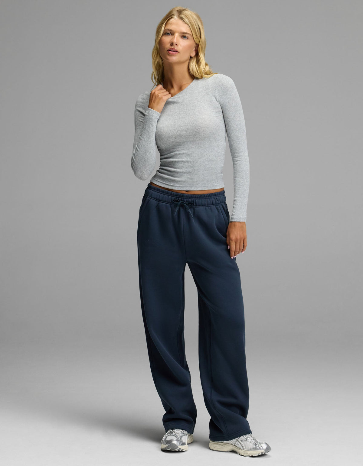 A woman with blonde hair and a neutral expression stands against a gray background, wearing the Cozy Earth Women’s Fine Ribbed Long Sleeve Tee in light gray, paired with loose navy sweatpants and silver sneakers.|Color:Heather Grey