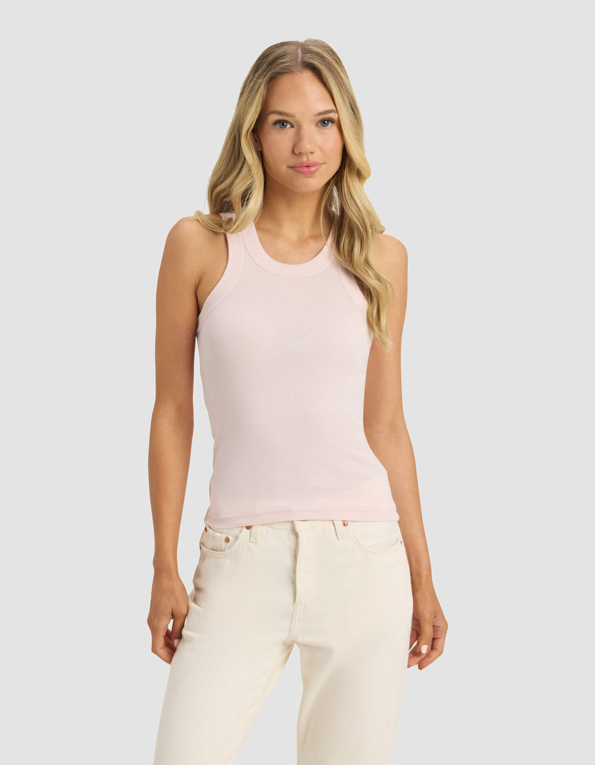 A woman with long blonde hair wears Cozy Earth's Women's Fine Ribbed Tank in light pink and off-white jeans, standing against a plain light gray background and facing forward with a neutral expression.|Color:Ballet