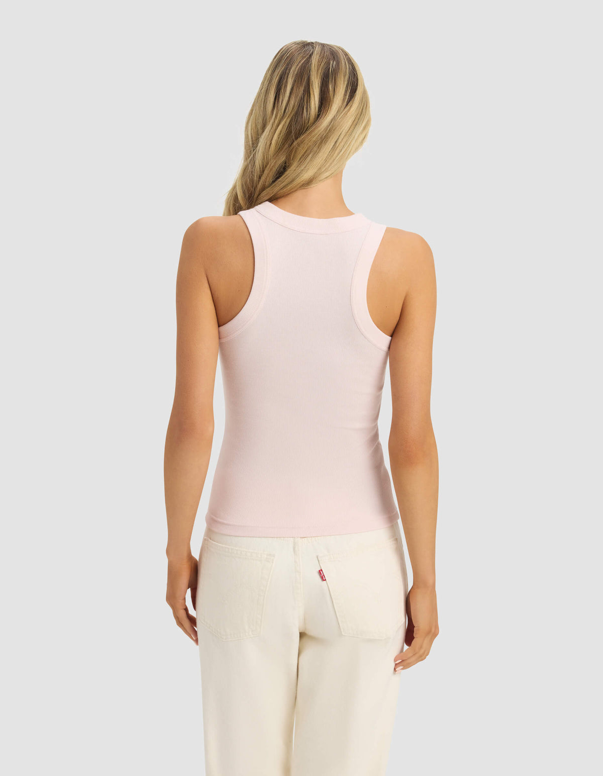 A woman with long blonde hair, seen from the back, wears the Cozy Earth Women's Fine Ribbed Tank in light pink and off-white pants, standing against a plain light gray background.|Color:Ballet