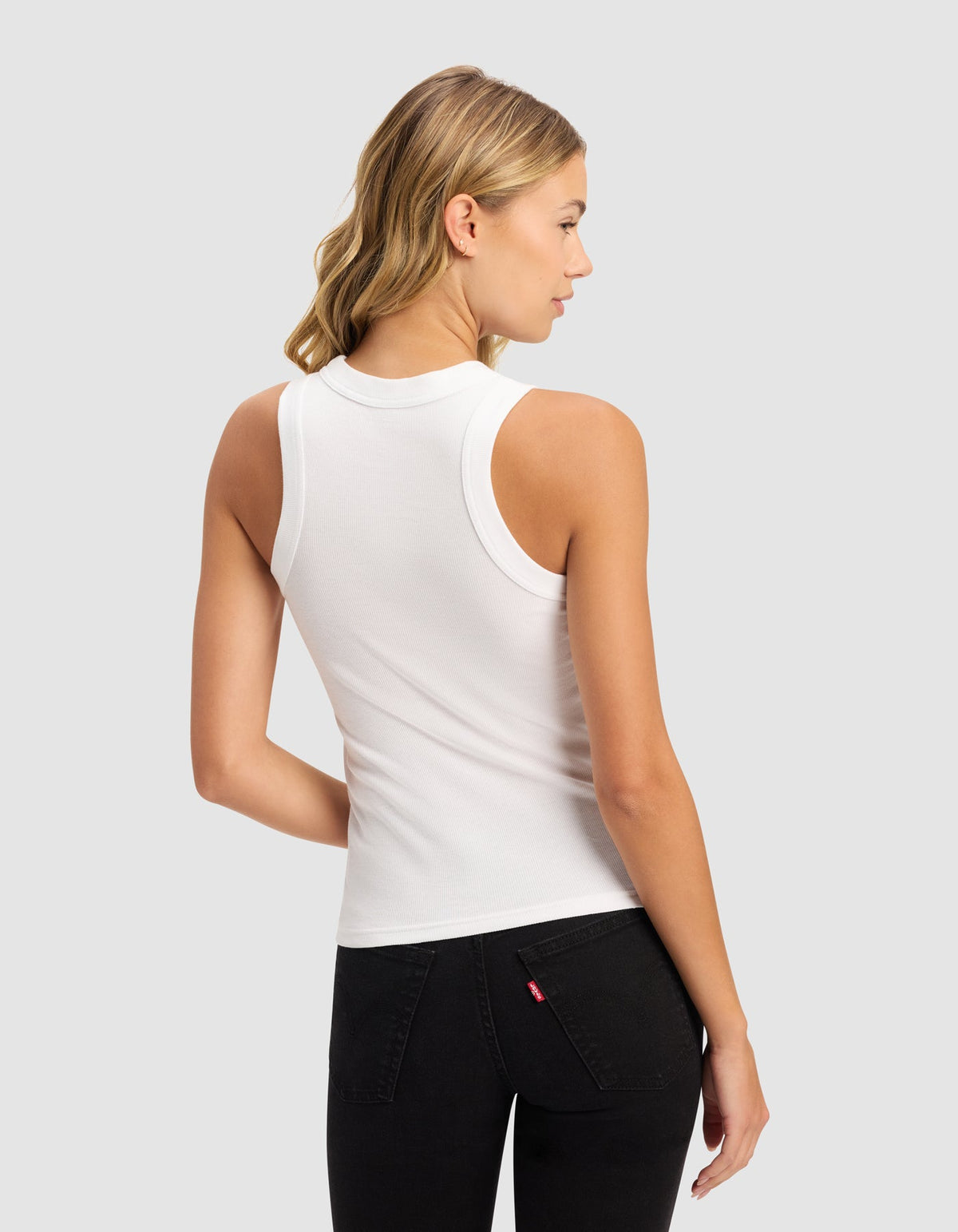 A woman with blonde wavy hair stands with her back to the camera, wearing Cozy Earth's Women's Fine Ribbed Tank in white and black jeans, set against a light gray background.|Color:White