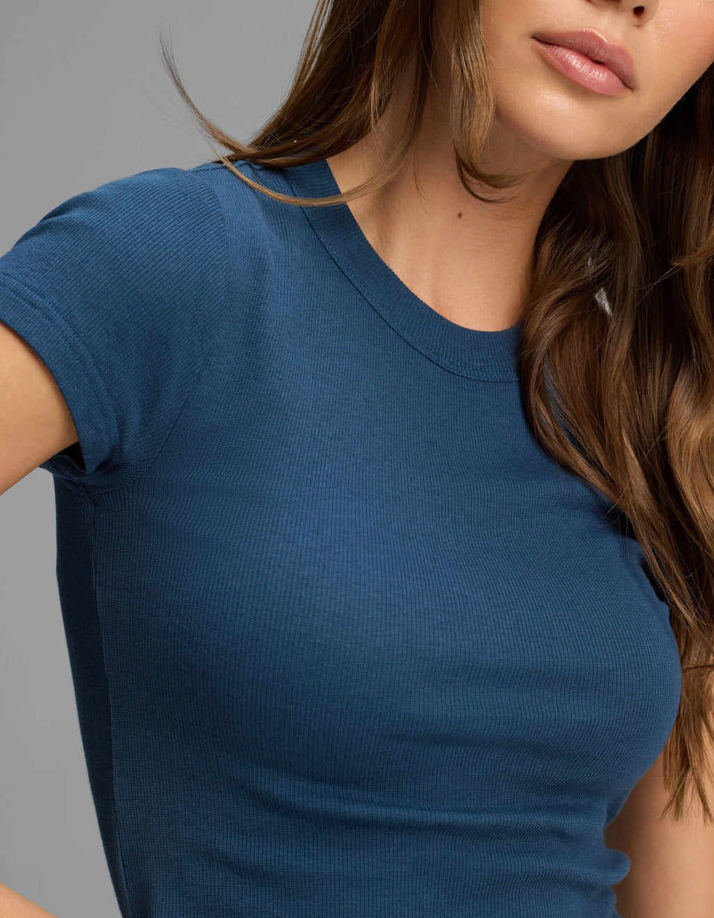A woman with long brown hair is shown from the torso up, modeling the Cozy Earth Women's Fine Ribbed Tee—a fitted, short-sleeve, blue ribbed t-shirt—against a plain gray background.|Color:Obsidian