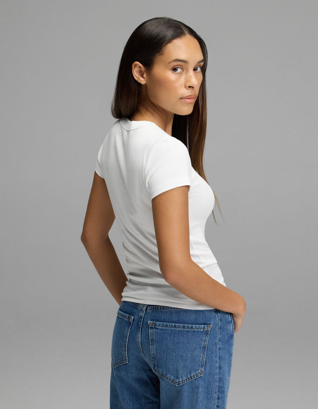 A woman with long brown hair wears the Cozy Earth Women’s Fine Ribbed Tee in white and blue jeans, standing sideways and looking over her shoulder against a plain gray background.|Color:White