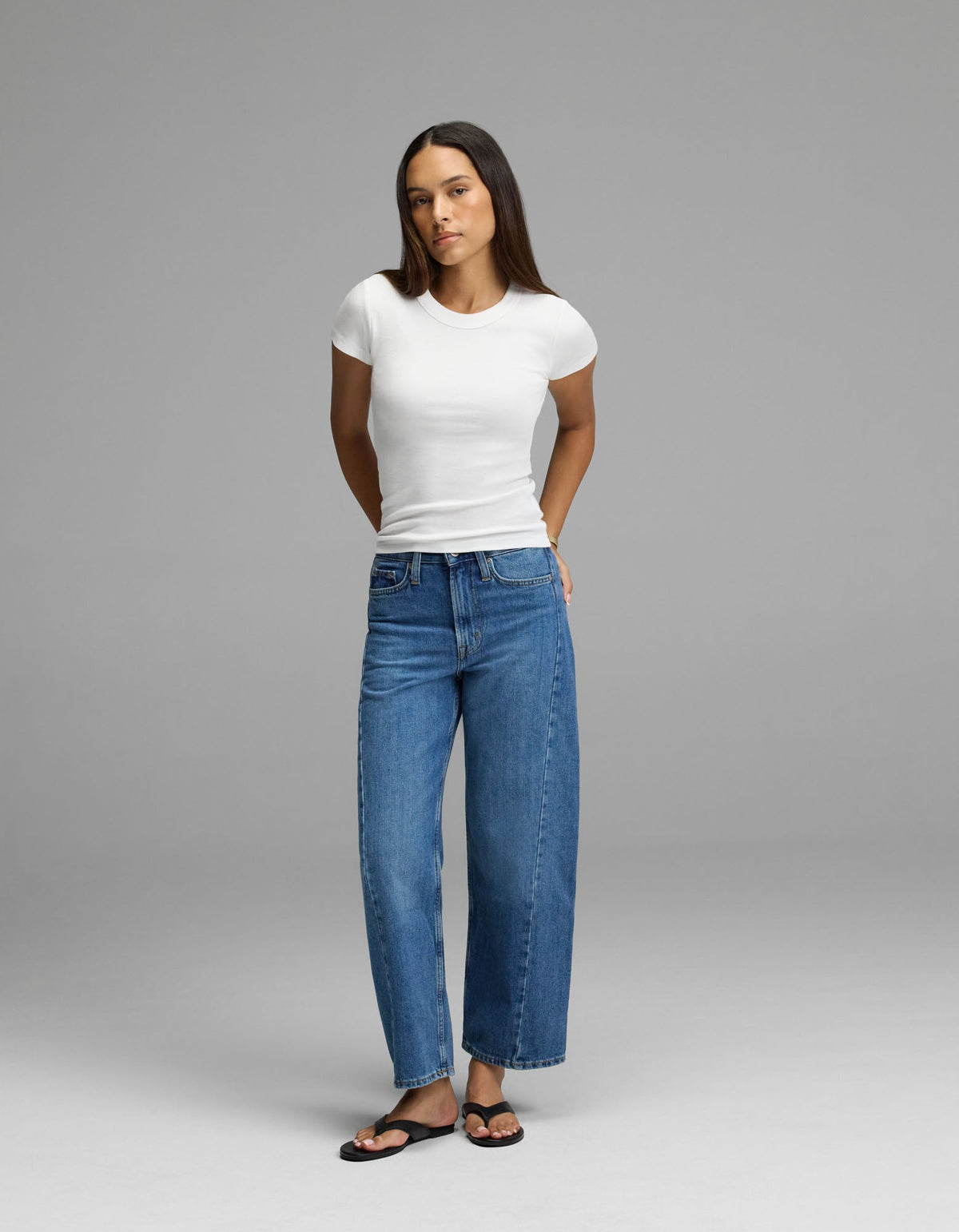 A woman stands against a plain gray background wearing the Cozy Earth Women’s Fine Ribbed Tee, styled with blue wide-leg jeans and black sandals. Her arms are behind her back, and she has long straight hair.|Color:White