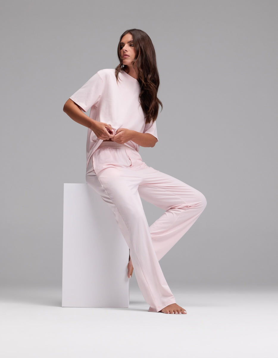 A woman with long brown hair wears the Cozy Earth Women’s First Layer Pajama Pant in light pink and sits barefoot on a white block against a plain gray background.|Color:Ballet