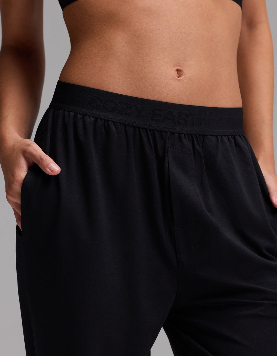 A person wearing Cozy Earth’s Women’s First Layer Pajama Pant in black, with hands in the pockets, paired with a black sports bra. The pants feature a waistband that displays “COZY EARTH,” and the background is plain and neutral.|Color:Jet Black